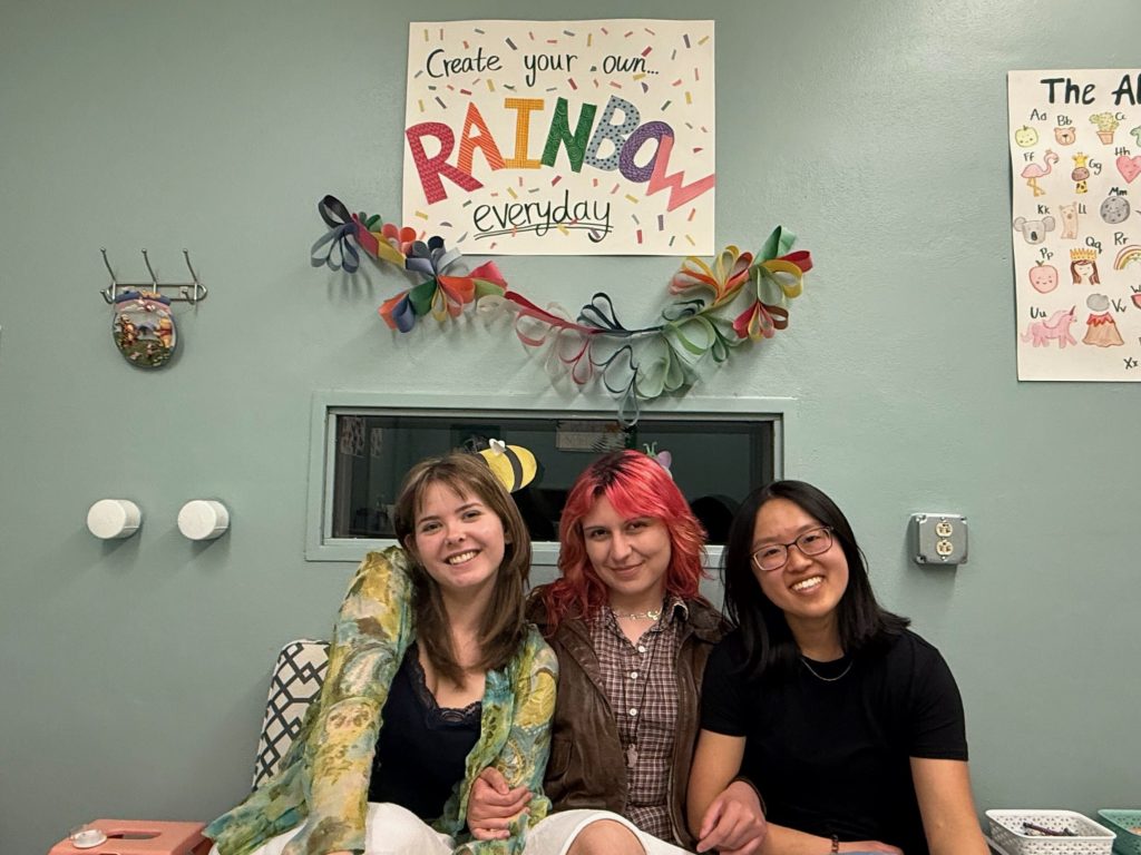 Three Social and Moral Learning Lab members sitting together and smiling beneath a colorful handmade ‘Create Your Own... Rainbow Everyday’ sign.