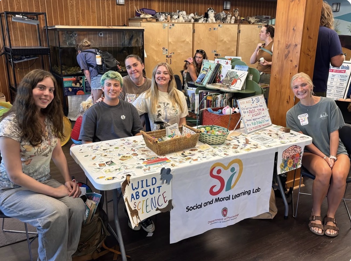 ocial and Moral Learning Lab members gathered around an outreach table at the Lussier Family Heritage Center, displaying research materials, toys, and colorful decorations.