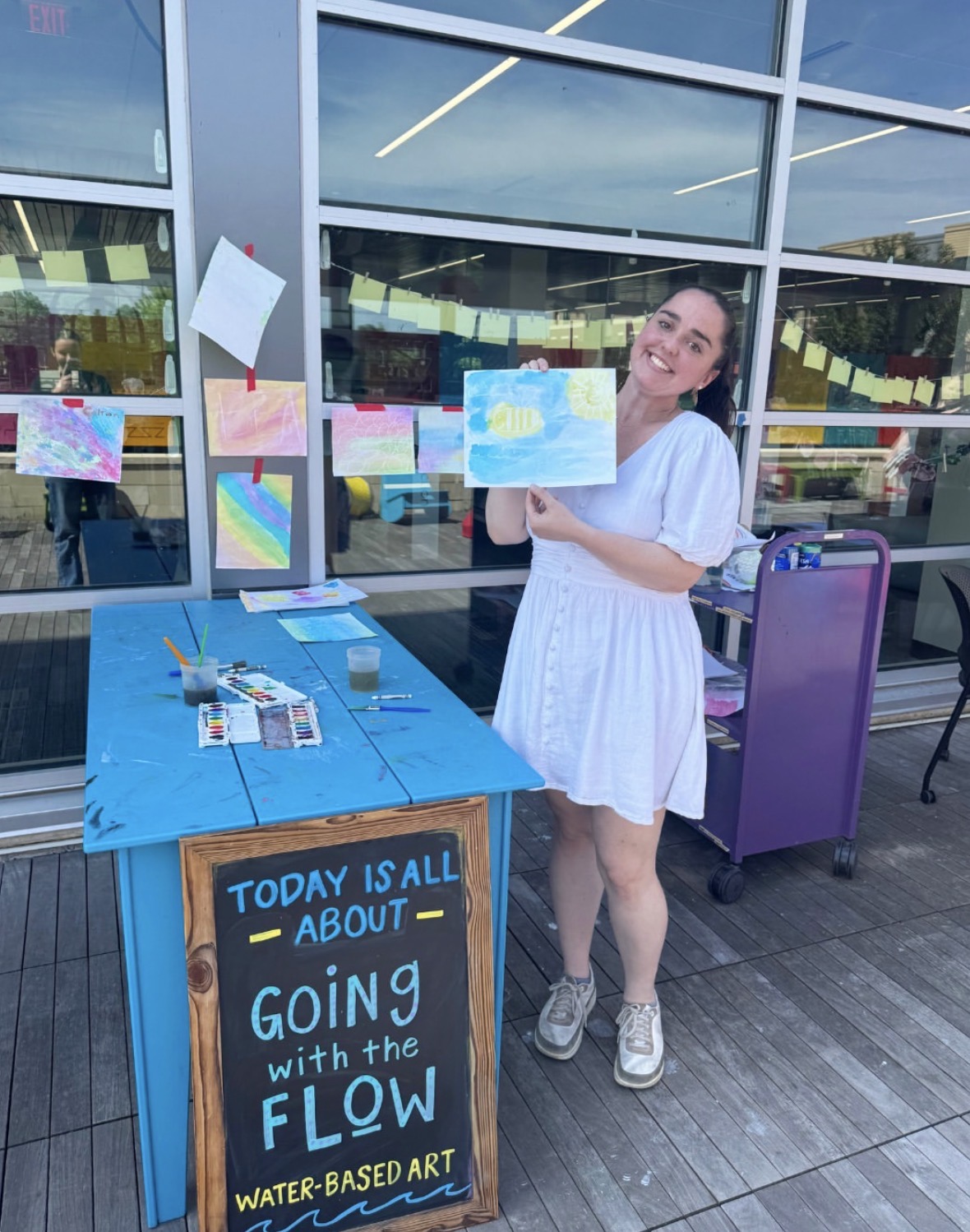 A Social and Moral Learning Lab member holding up a colorful watercolor painting at Pinney Library, with additional artwork and painting supplies displayed on a table.