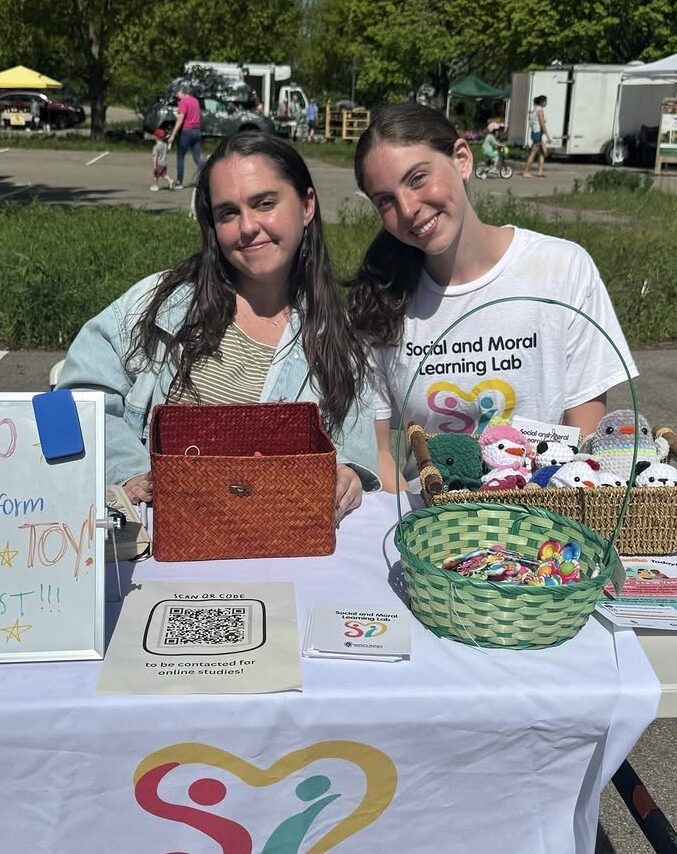 Two Social and Moral Learning Lab members standing behind an outreach table at a Madison farmers' market, displaying research information, colorful toys, and recruitment materials.