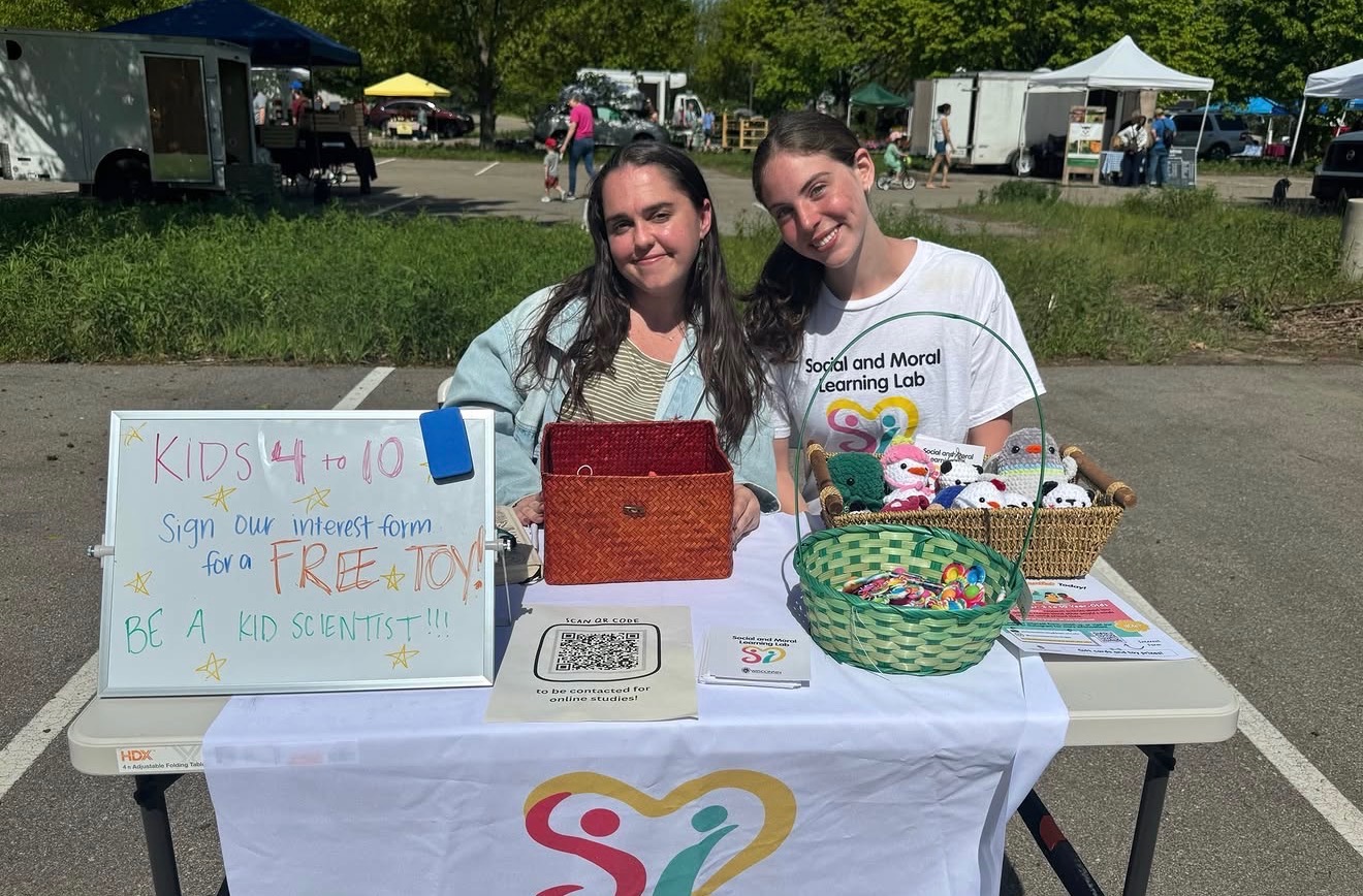 Lab manager Jane and research assistant Kate smile behind a Social and Moral Learning Lab outreach table at a Madison farmers' market, displaying study information and baskets of toys for children.