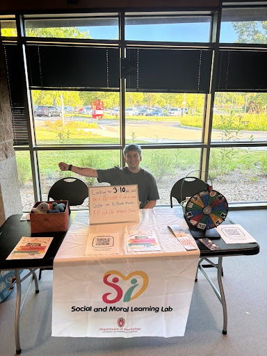 Social and Moral Learning Lab member smiles behind an outreach table at Family Fun Night at Warner Park Community Recreation Center, displaying research information, a colorful prize wheel, and baskets of toys for children.