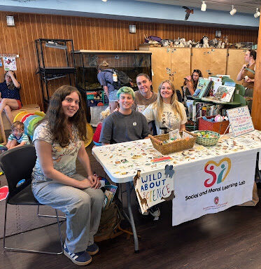 Social and Moral Learning Lab members and lab manager smiling together behind their community outreach table at the Lussier Family Heritage Center, featuring research materials, colorful toys, and handmade decorations.