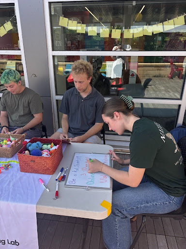 Three Social and Moral Learning Lab members preparing for a community outreach event at Pinney Library, arranging colorful toys and creating a handwritten sign at their table.