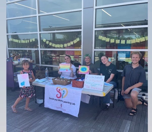 Three Social and Moral Learning Lab members display colorful watercolor paintings created using bubbles at a community outreach event at Pinney Library, seated behind a table with art supplies and research information.
