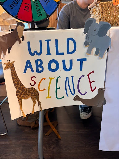 Handmade ‘Wild About Science’ sign created by a Social and Moral Learning Lab research assistant, featuring colorful lettering and animal illustrations, including a giraffe, elephant, goat, and seal, displayed at a community outreach table.